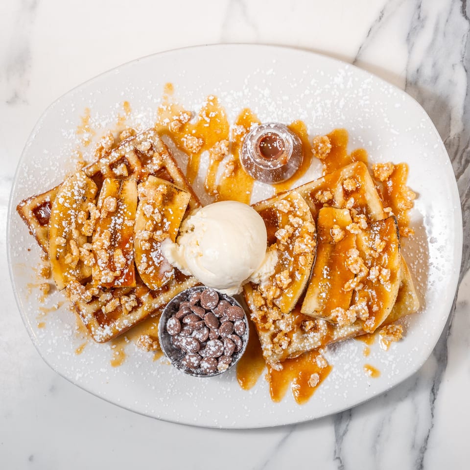 Waffles topped with caramelized bananas, ice cream, chocolate chips, and syrup on a white plate.