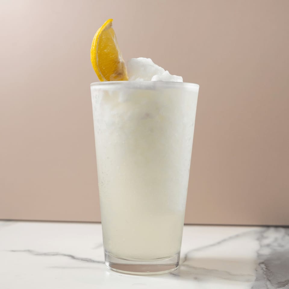 A frosty glass of frozen lemonade, garnished with a fresh lemon wedge, on a marble surface.