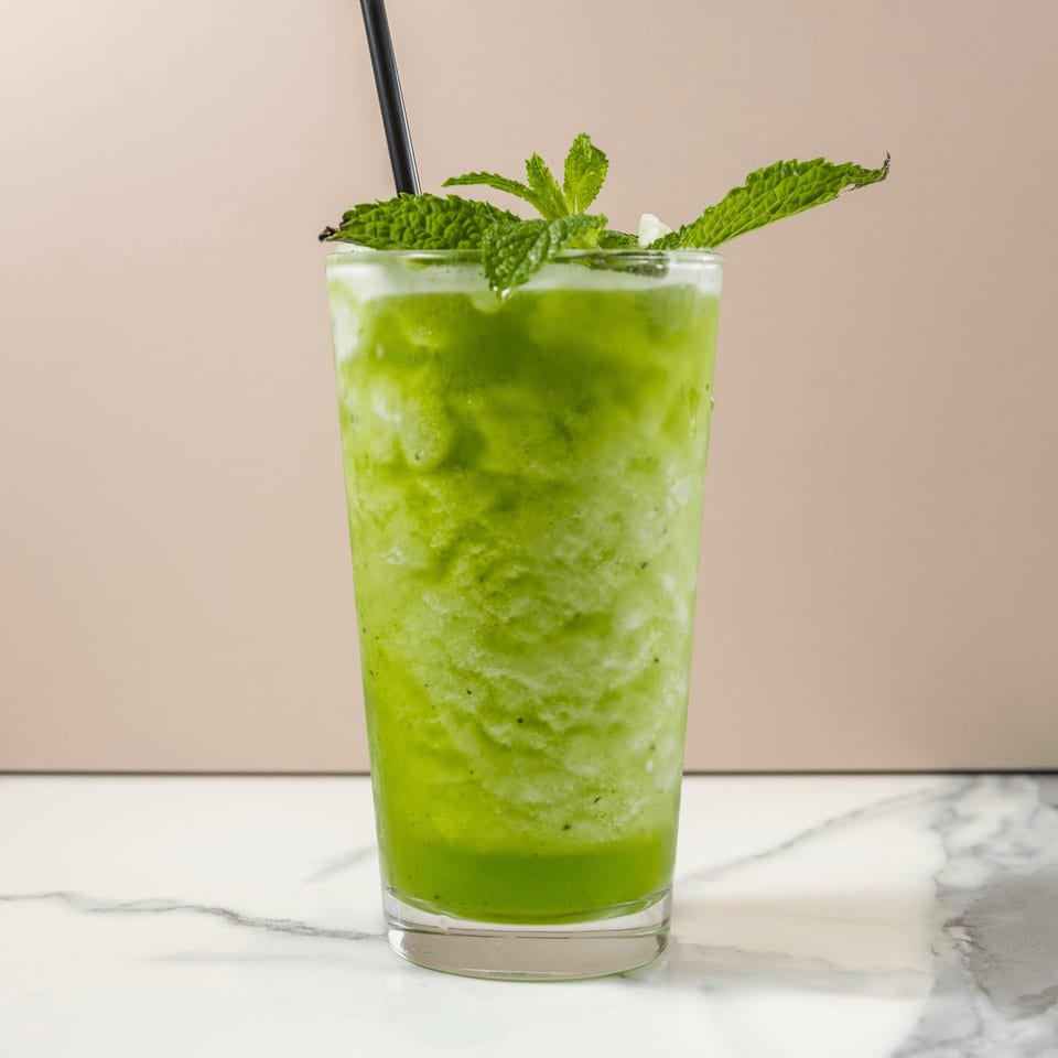 A vibrant green smoothie with mint leaves and a black straw, on a marble surface.