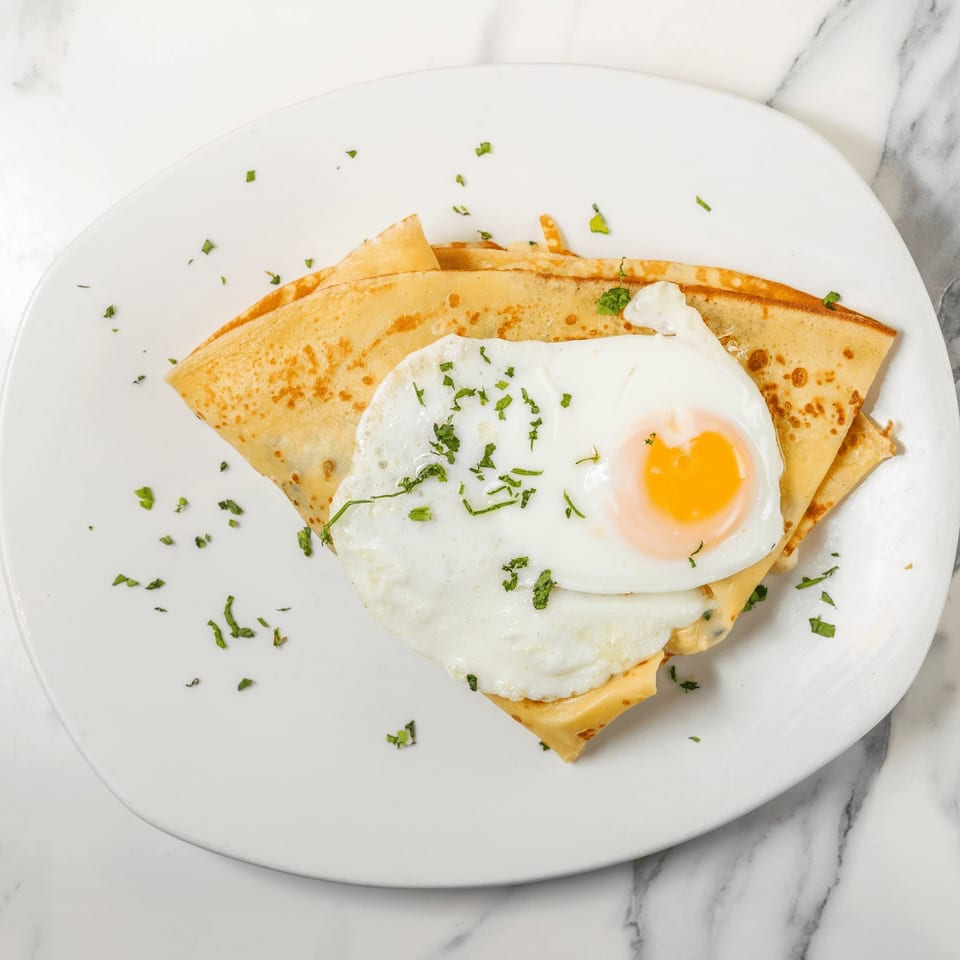 Savory crepe topped with a fried egg, melted cheese, and fresh herbs on a white plate.