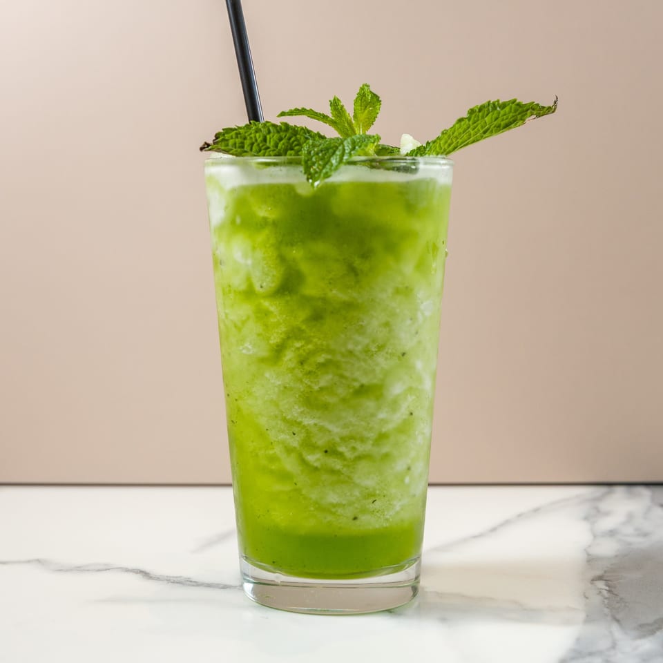 Green smoothie with mint leaves and a black straw in a tall glass on a marble surface.